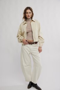 Beige leichte Jacke mit elastischen Bündchen über einem gestreiften Langarmshirt. Weiße Hose mit weitem Bein und schwarze Stiefeletten vervollständigen das Outfit.