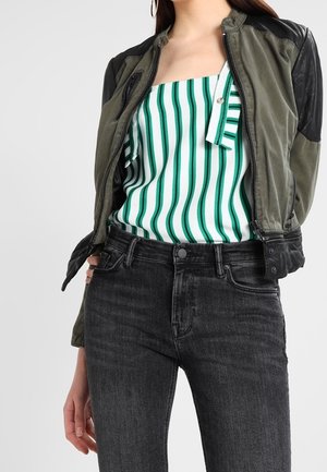 Femme portant une veste verte et noire par-dessus un haut blanc à rayures verticales vertes et un jean noir taille haute.