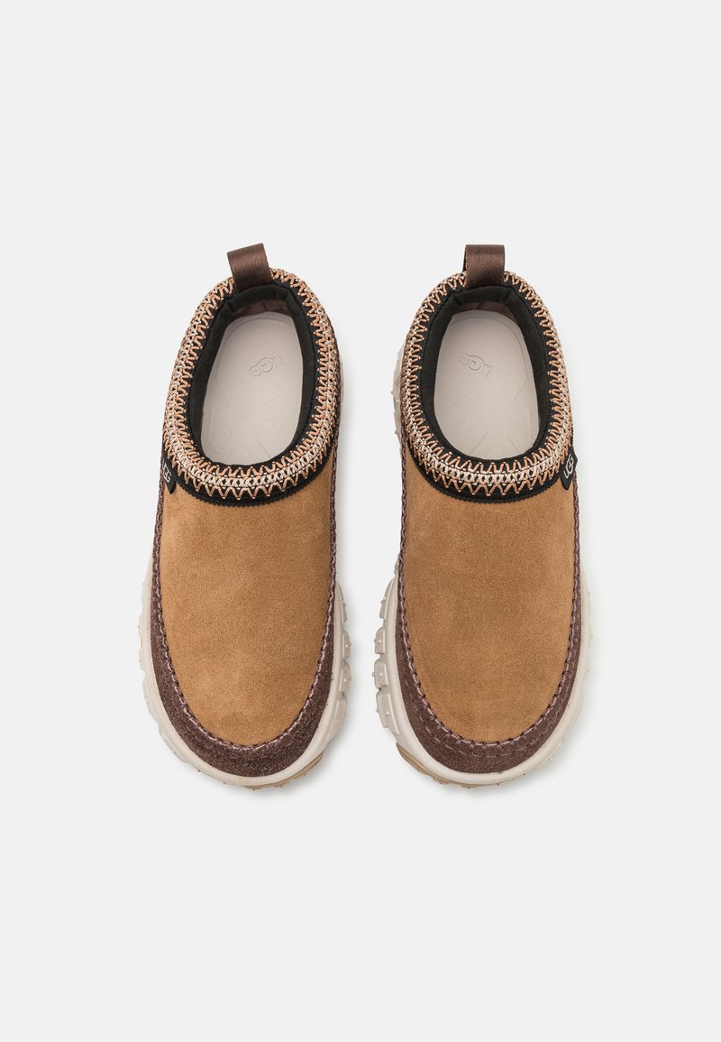 Zapatos de ante marrón tipo slip-on con una suela de goma texturizada, acentos en marrón oscuro en contraste y un cuello decorativo con costuras.