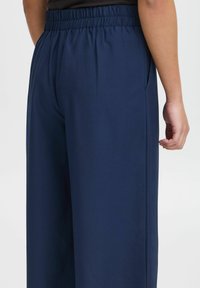 Navy blue, wide-leg trousers with an elastic waistband, featuring side pockets and a smooth texture, shown from the back.