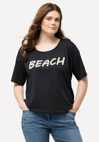 Sort kortærmet T-shirt med "BEACH" trykt i hvid, scoop neck design, lavet af blødt stof, parret med blå jeans.