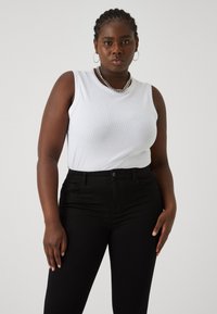 Vit ribbad ärmlös topp i kombination med svarta jeans. Toppens har en rund halsringning, och modellen bär ett silverfärgat kedjearmband och hoopsörhängen.