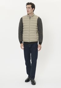 Beige vadderad väst med hög krage över en mönstrad mörk tröja, tillsammans med mörka jeans i denim och svarta skor. Enkelt mönster, åtsittande silhuett.