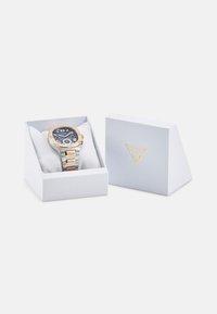 Montre Guess avec un cadran bleu marine, des accents en or rose et un bracelet en acier inoxydable, présentée dans une boîte blanche avec le logo de la marque.