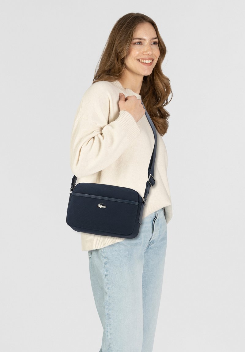Donna sorridente che indossa un maglione crema e jeans chiari, con una borsa a tracolla blu navy con logo argentato sulla tracolla, portata sulla spalla.