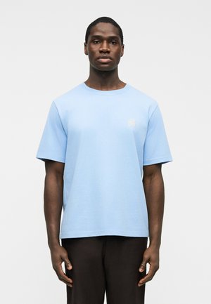 Ung mann som står vendt fremover, iført en lyseblå kortermet T-skjorte med et lite hvitt kløveremblem på brystet og mørke bukser.