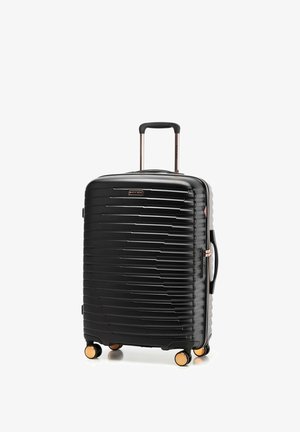 Valise rigide noire à coque dure avec rainures horizontales, poignée extensible, poignée latérale et quatre roues pivotantes avec accents dorés sur fond blanc.