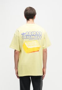 Κίτρινη oversized μπλούζα με κοντό μανίκι που διαθέτει μπλε κείμενο "ADIDAS ORIGINALS" και μια γραφική απεικόνιση ενός μπλοκ βουτύρου στην πλάτη; μαλακή υφή.