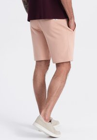Světle růžové bavlněné kraťasy s bočními kapsami, se elastickým pasem. Nosič je v kombinaci s béžovými teniskami, které mají minimalistický design.