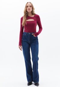 Tummanpunainen pitkähihaisella puserolla, jossa on leikkauskohta, yhdistetty korkeavyötäröisiin tummansinisiin farkkuleggingeihin, joissa on etutaskut ja vyölenkit.