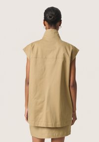 Beige mouwloze shirt met een hoge kraag, gemaakt van een zachte stof. Heeft een eenvoudig, rechtlijnig ontwerp en een rechte zoom aan de achterkant.