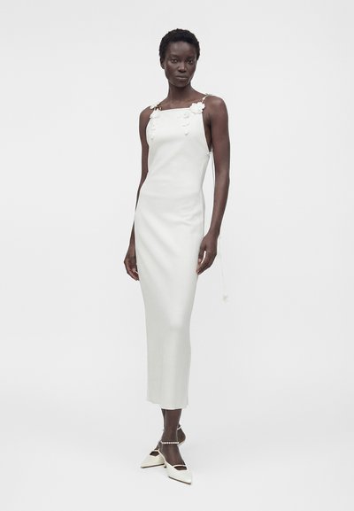 Γυναίκα που φορά λευκό midi φόρεμα χωρίς μανίκια με floral τιράντες, συνδυασμένο με λευκά μυτερά παπούτσια και βραχιόλι αστραγάλου με μαργαριτάρια, στέκεται σε απλό φόντο.