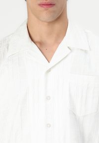 Camisa blanca de manga corta que presenta una tela texturizada, un collar tipo esparcido, un bolsillo frontal y cuatro botones en la tapeta.