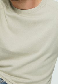 Hellbeiger Strickpullover mit Rundhalsausschnitt und gerippten Bündchen. Der Stoff wirkt glatt und weich mit minimaler Textur.