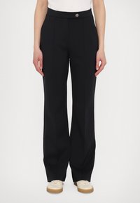 Black trousers with a smooth texture, flared design, and a front button closure. Paired with light-colored shoes, featuring a rubber sole.