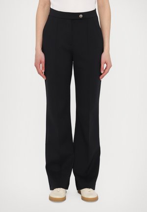 Pantalones negros con una textura suave, de diseño acampanado y cierre de botón en la parte delantera. Combinados con zapatos de color claro, con suela de goma.