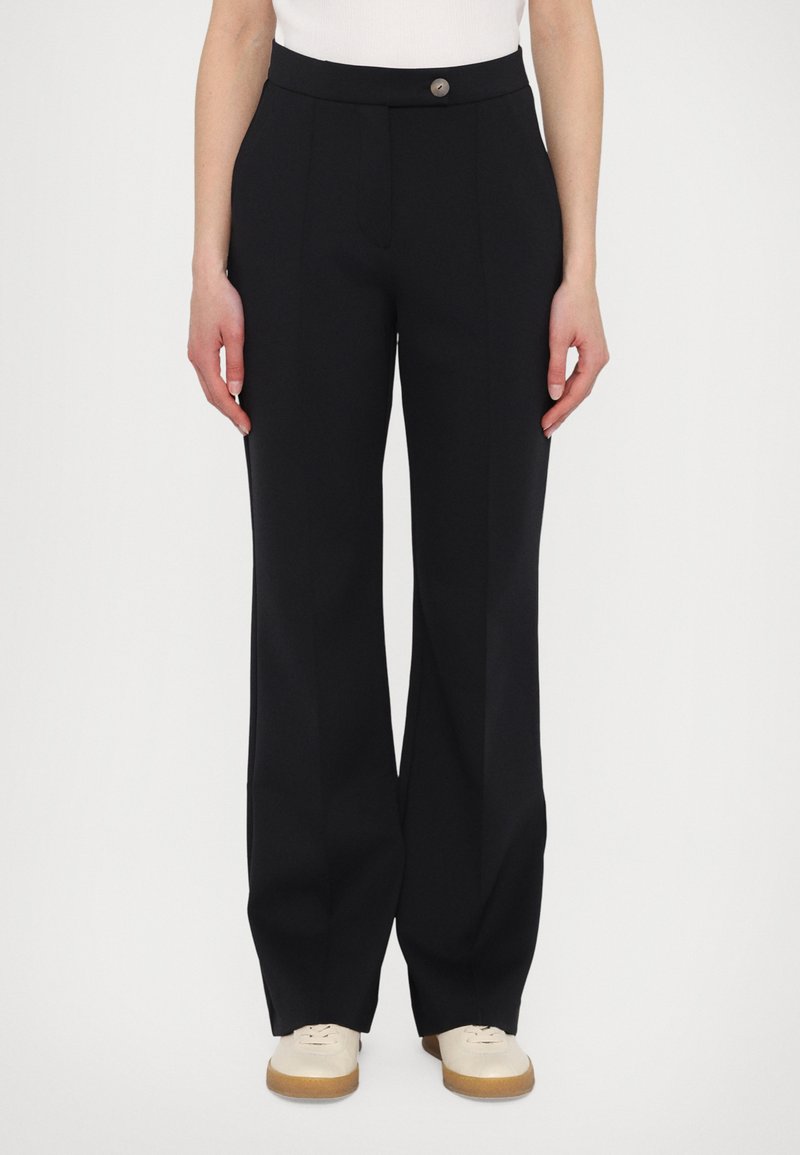 Black trousers with a smooth texture, flared design, and a front button closure. Paired with light-colored shoes, featuring a rubber sole.