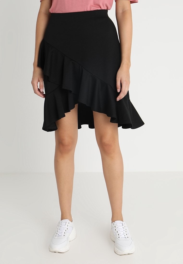 Gonna nera asimmetrica con orlo a balze, realizzata in tessuto testurizzato. Indossata con un top rosa e sneakers bianche, che mostrano un design a strati.