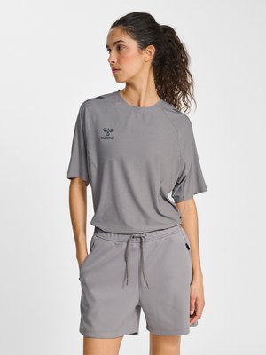 Kobieta z kręconymi włosami, w szarej koszulce z krótkim rękawem i pasujących krótkich spodenkach, z rękami w kieszeniach, patrząca w lewo.