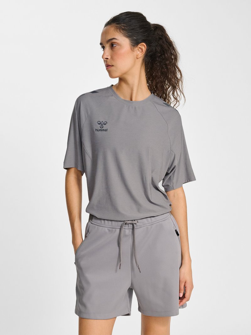 Kvinde med krøllet hår iført en grå t-shirt med korte ærmer og matchende shorts med hænderne i lommerne, kiggende mod venstre.