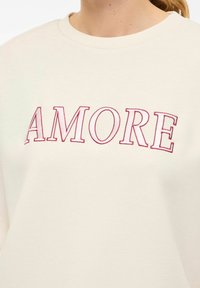 Sweatshirt beige clair avec un col rond et le mot "AMORE" en broderie rose audacieuse sur la poitrine. Texture de tissu lisse.