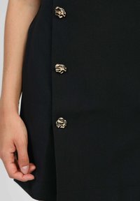 Main reposée près de l'ourlet d'un vêtement noir avec trois boutons métalliques de forme irrégulière alignés verticalement.