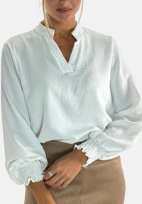 Blouse blanche avec un col en V et des manches longues bouffantes. Dispose de poignets froncés et d'un tissu texturé. Associée à une jupe beige.