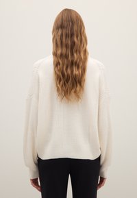 Cremefarbener, gerippter Pullover mit lockerem Schnitt, weiten Ärmeln und strukturierter Oberfläche. Von hinten gesehen über einer schwarzen Hose getragen.