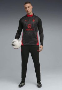 Musta ja punainen urheiluasu, jossa on vetoketjullinen takki ja siihen sopivat housut. Takissa on teksturoitu muotoilu ja logo, ja se pitää valkoista jalkapalloa.