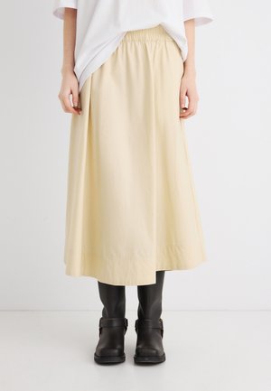 Osoba ubrana w beżową spódnicę midi z elastycznym pasem, biały top oraz czarne skórzane botki na kostkę z paskami i klamrami.