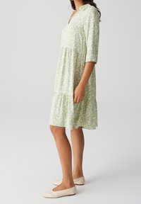Robe vert clair avec motif abstrait blanc, décolleté en V, manches trois-quarts, ourlet superposé et tissu doux. Portée avec des chaussures plates beiges.