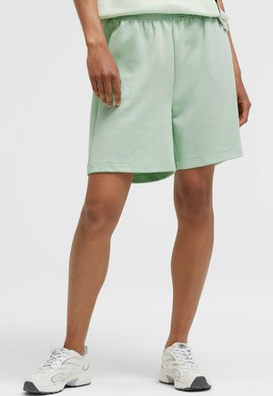 Legs of a person wearing light green knee-length shorts and white sneakers standing on a plain background.