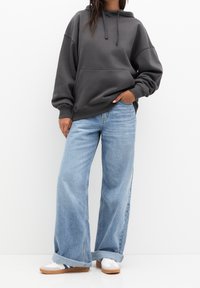 Sudadera gris oscuro con un bolsillo delantero, corte oversized, combinada con jeans de pierna ancha de color azul claro y zapatillas blancas con suelas marrones y rayas azules.