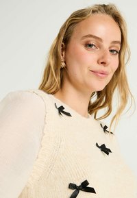 Pull en maille crème avec un motif texturé, mettant en valeur des accents en ruban noir à l'avant. Modèle portant un tissu léger et transparent en dessous.