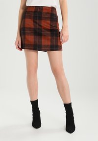 Donna vestita con una mini gonna a quadri marrone e nera, top bianco e stivali neri alla caviglia, in piedi su uno sfondo bianco semplice.