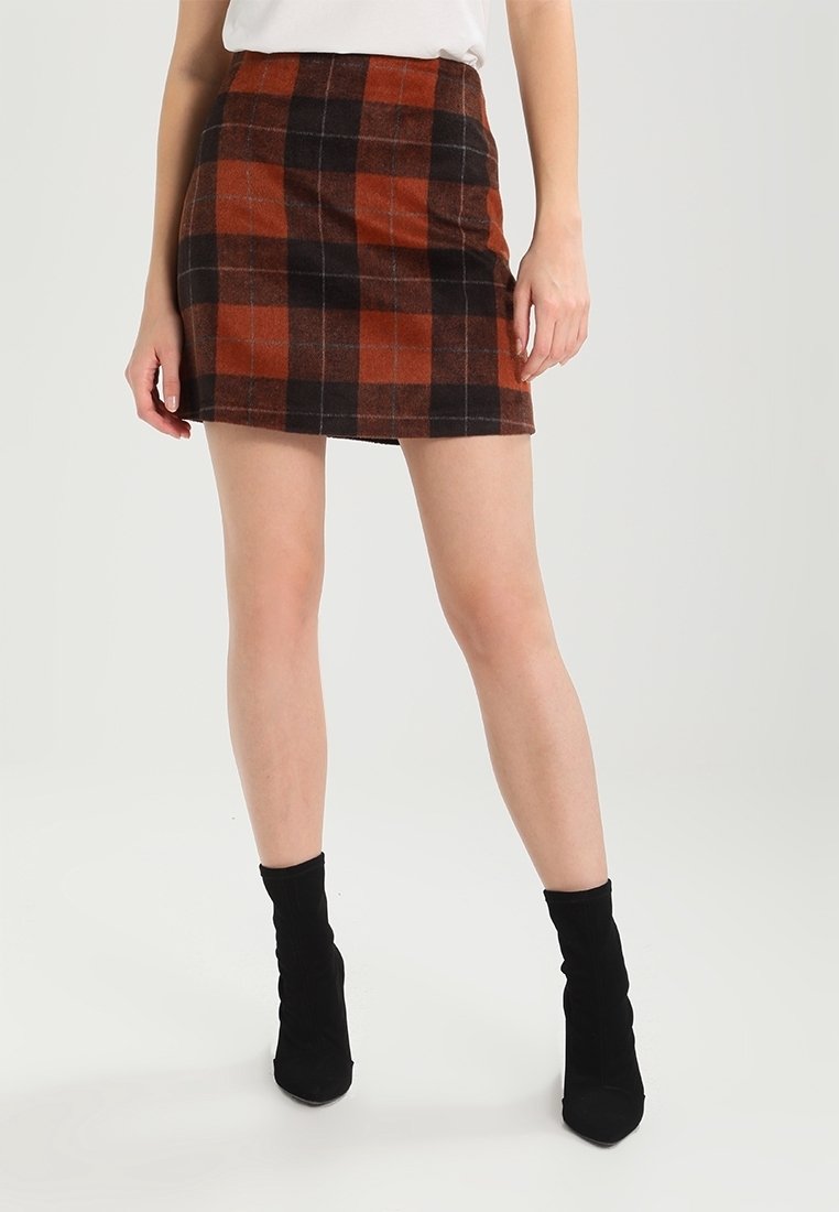 Donna vestita con una mini gonna a quadri marrone e nera, top bianco e stivali neri alla caviglia, in piedi su uno sfondo bianco semplice.