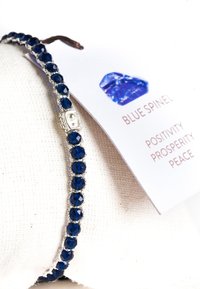 Blauer Spinellarmband mit facettierten blauen Steinen, silbernen Akzenten und geflochtenem Draht. Begleitet von einem Etikett, das Positivität, Wohlstand und Frieden beschreibt.