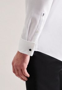 Camicia bianca con finitura testurizzata, caratterizzata da un bottone nero e un dettaglio floreale ricamato sul polsino. Vestibilità aderente al polso.