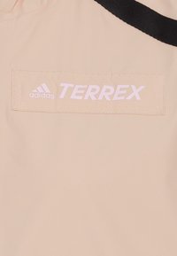 Terrex-jacka i mjuk beige tyg med en slät textur. Har en framträdande vit adidas-logotyp och sömnadsdetaljer på bröstfickan.