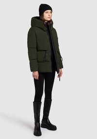 Groene pufferjas met een capuchon, met een vierkante vorm, ritssluiting en trekkoordaccenten. Gecombineerd met zwarte leggings en kniehoge laarzen.