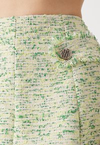 Tissu en tweed vert et crème avec un tissage texturé, présentant un bouton rond en métal doré avec logo et des bords effilochés pour une touche supplémentaire.