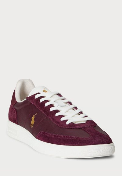 Sneaker bordeaux in camoscio e pelle, con lacci bianchi, suola texturizzata e un accento logo dorato. Design classico a bassa altezza.