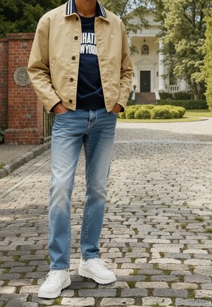 Mand iført beige jakke, marineblå grafisk sweatshirt, lyseblå jeans og hvide sneakers, der står på en brostensbelagt sti foran et stort hus.