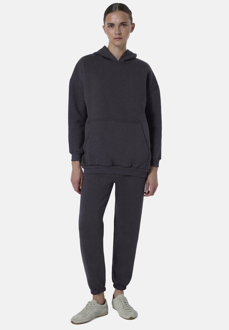 Donkergrijze hoodie met een zak en een losse pasvorm, gecombineerd met bijpassende joggingbroeken met elastische manchetten, gemaakt van zachte stof. Sneakers maken de outfit compleet.