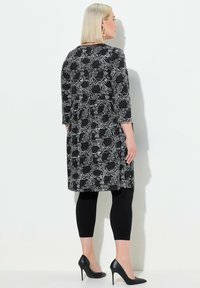 Schwarz-weiß gemustertes Kleid mit Dreiviertelärmeln, Rundhalsausschnitt und lockerer Passform, kombiniert mit schwarzen Leggings und hohen Schuhen.