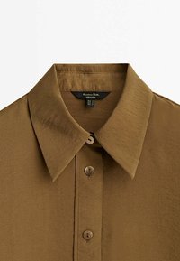 Chemise brune à col avec une texture lisse, fermeture à boutons et détails de couture visibles. Étiquette visible à l'intérieur du col.