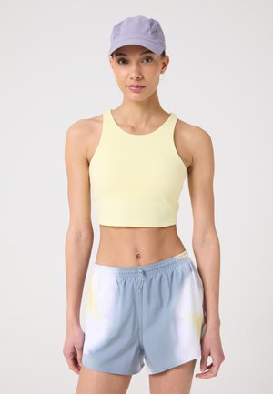 Woman wearing a lavender cap, pale yellow cropped tank top, and light blue and white athletic shorts against a plain background.