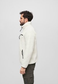 Polarowy pulower w kolorze off-white, z wysokim kołnierzem, kieszenią na zamek na piersi, regulowanymi sznurkami, teksturowana tkanina, zestawiony z ciemnymi spodniami cargo.