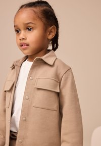 Veste beige boutonnée avec deux poches avant, en tissu doux. L'enfant porte une chemise blanche en dessous, au design simple.