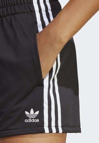 Handen vilar i fickan på svarta Adidas shorts med vita ränder och logotyp på sidan.